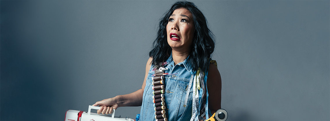 Kristina Wong posing against a dark backdrop. She is holding a sewing machine and miming being scared.