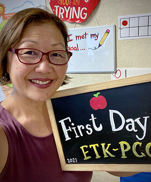 A photo of Jane in a classroom smiling at the camera and holding a chalkboard sign that has an apple and the words &quot;First Day&quot; written on it
