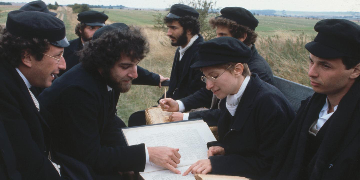Still from the movie showing Mandy Patinkin and Barbara Streisand sitting with other men in the back of a truck reading religious text