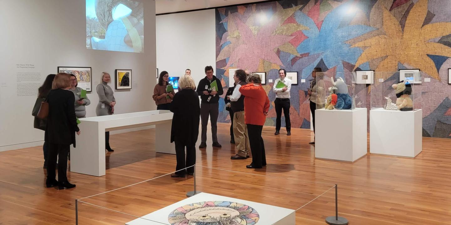 A gallery space filled with illustrations, artwork on pedestals, and a video presentation. People walk throughout and look at the art.