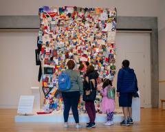 Photo of a gallery space with a large quilt hanging from the ceiling. Three adults and a child stand with their backs to the camera looking at the quilt together.