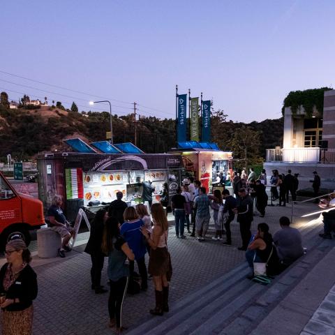 Photo of the front steps of the Skirball. People are standing around talking and ordering food from parked trucks. The words "Late Night!" are projected in lights on the side of the building.