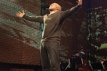 Photo of Bryonn Bain performing on stage. He is standing with his arms outstretched and facing upward. Behind him, a group of musicians can be seen with a screen.