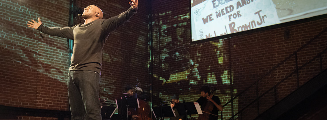 Photo of Bryonn Bain performing on stage. He is standing with his arms outstretched and facing upward. Behind him, a group of musicians can be seen with a screen.