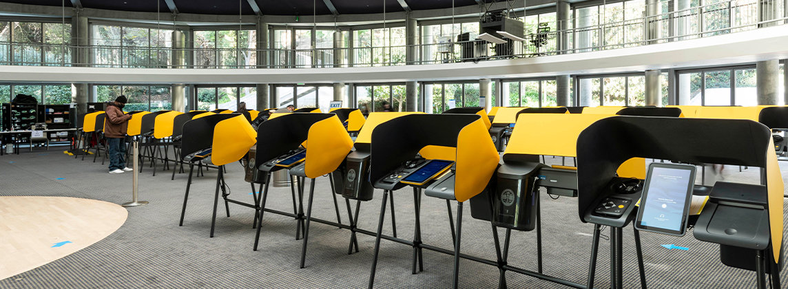 Photo of a large indoor voting center. A series of yellow and black voting machines are lined up in curved rows.