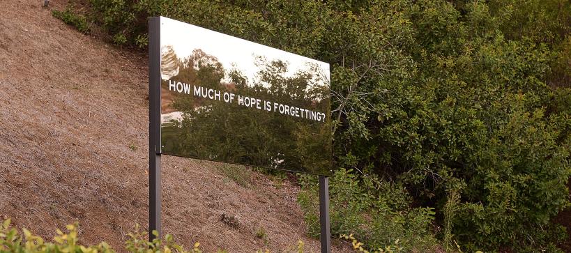 A large reflective billboard on a hillside that says, "How much hope is forgetting?". An outdoor artwork by Chloe Bass.