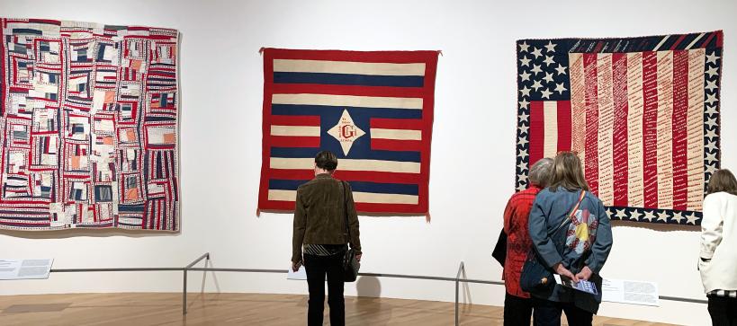 Four visitors in the gallery looking at three quilts with American flag designs in red, white and blue.