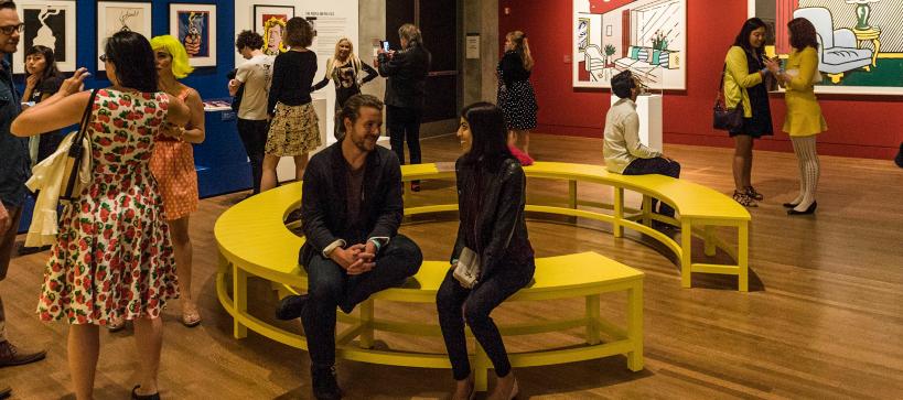 A man and women seated on a yellow circular bench in the middle of a gallery with people walking about