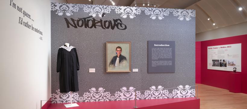 photo of the installation in the exhibition for Notorious RBG. It has a supreme court robe next to a portrait of Ruth Bater Ginsberg.