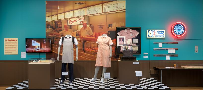 an exhibit of a deli setting with a checkerboard floor and 2 mannequins with deli uniforms on. A large photo behind of a deli counter.