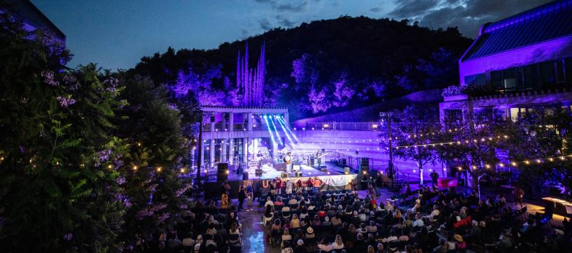 Photo of an outdoor concert at the Skirball. A large group of people are in the courtyard dancing to a band on stage. Lights are seen in the trees, strung across the courtyard, and from the stage.