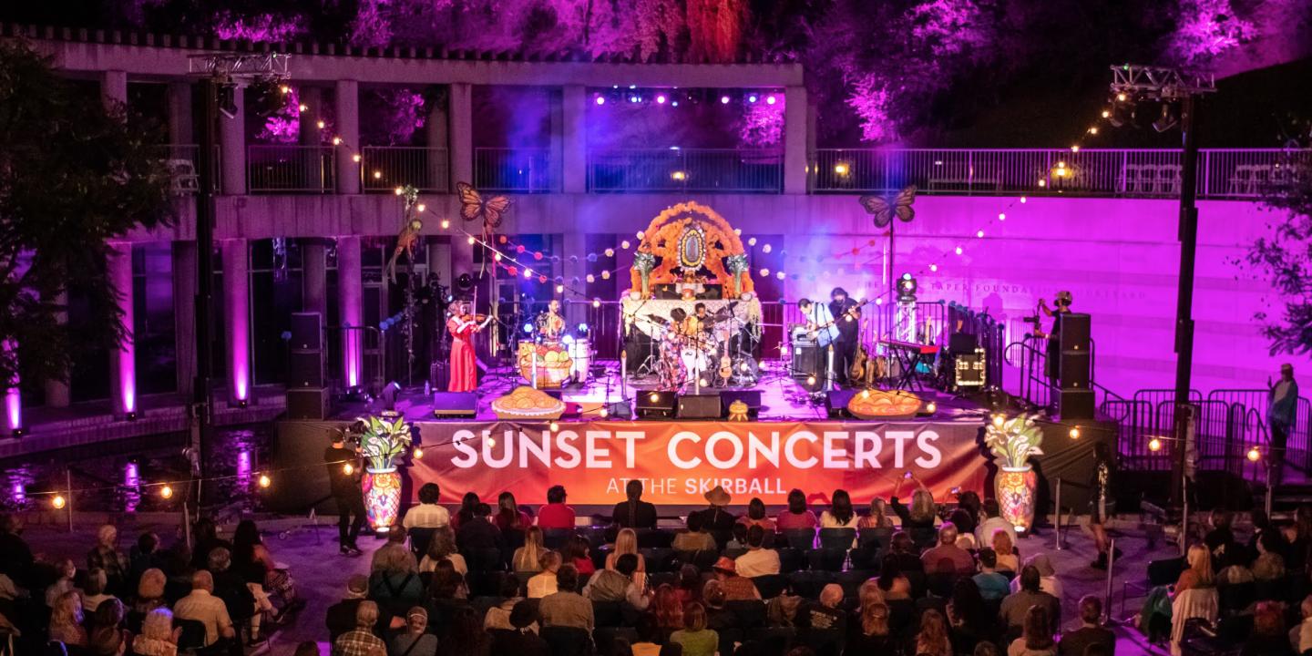 Image of outdoor stage with band playing and audience seated and listening. A large banner on the stage reads Skirball Stages