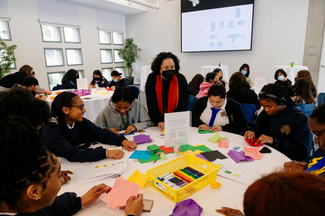 Photo of a group of students sitting around a white, round table folding colorful papers.