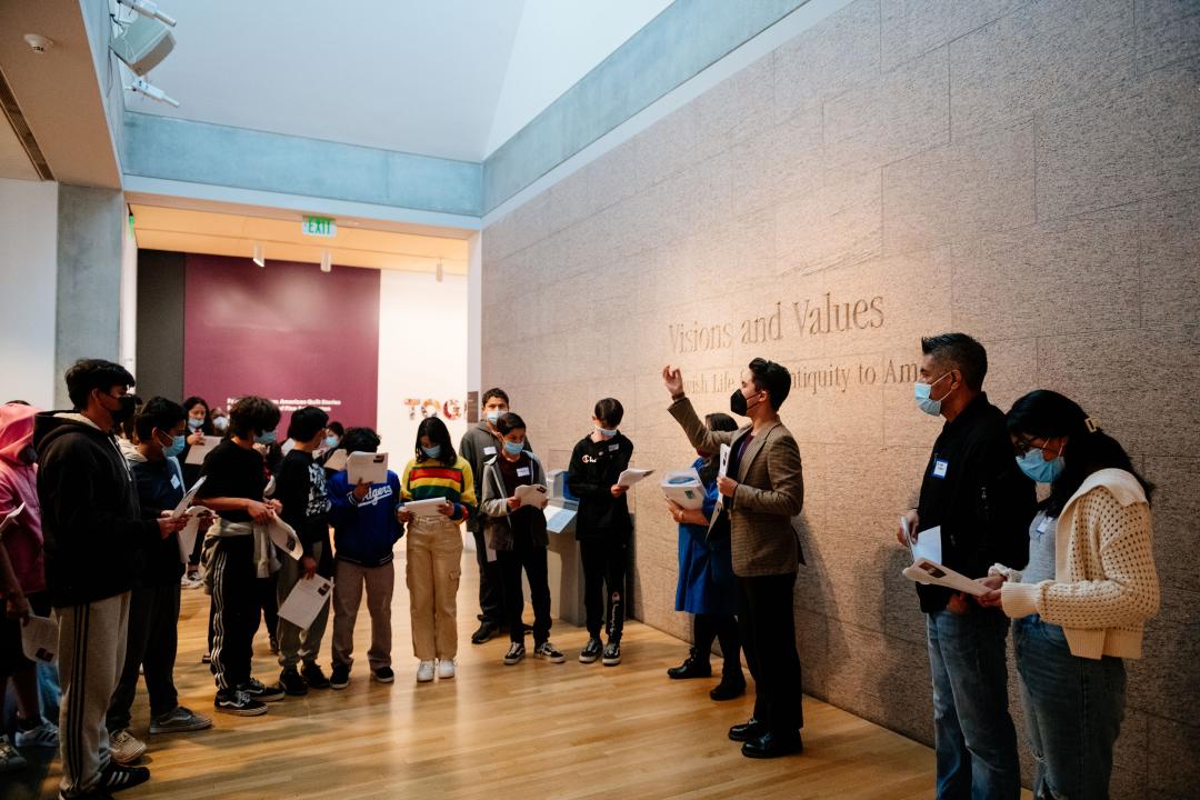 Photo of a group of students gathered around the entrance to the exhibition Visions and Values. An adult is seen gesturing to the students to orient them to the gallery.