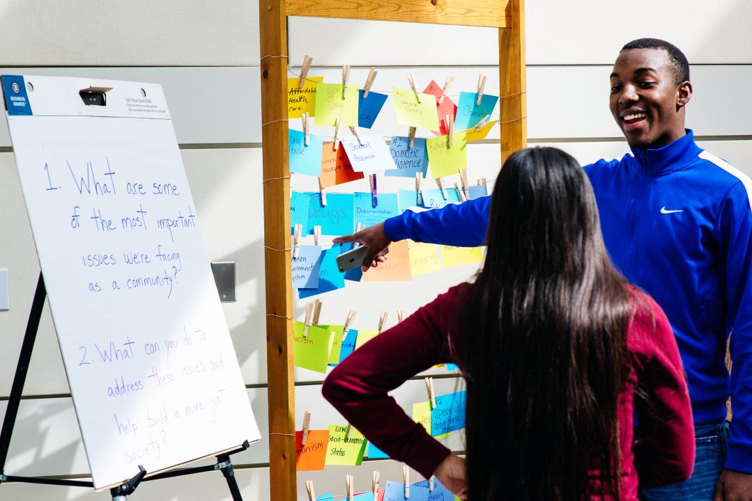 Two students talking and pointing at post-its and presentation boards with words and ideas on them.