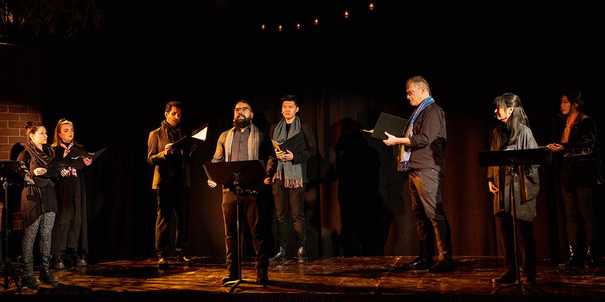 The group theatre dybbuk performing on a stage. Eight people stand reading from black binders on a dark stage wearing all black clothing except for colorful scarves around their necks.