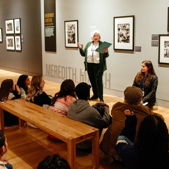 A docent standing in front of a photo in a gallery to a seated audience of teens.