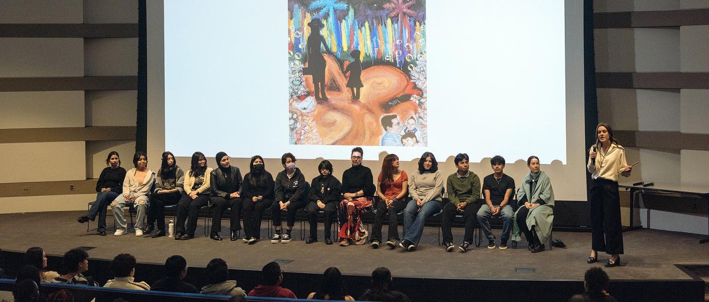 An auditorium watching a discussion on stage with a colorful piece of art on a projector behind them.