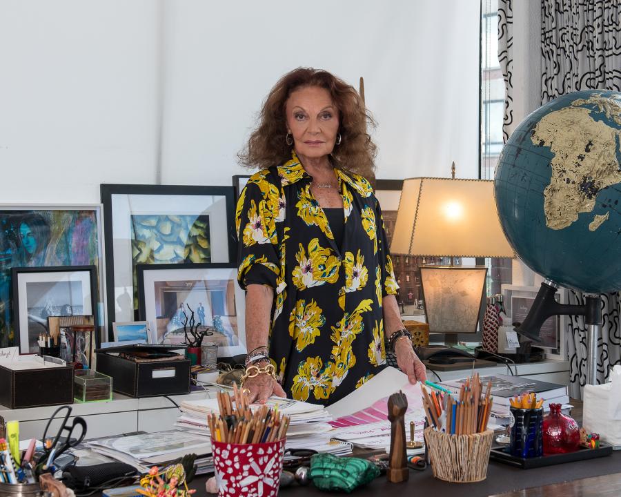 A woman with brown hair wearing a black and yellow dress stands behind a desk