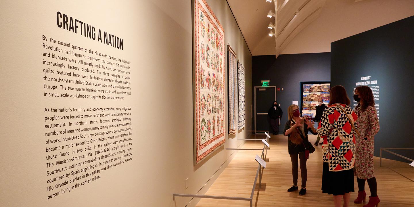 Image from inside a gallery space in Fabric of a Nation: American Quilt Stories. Large quilts are hanging on all walls and three women stand in the center of the frame discussing a quilt in front of them.