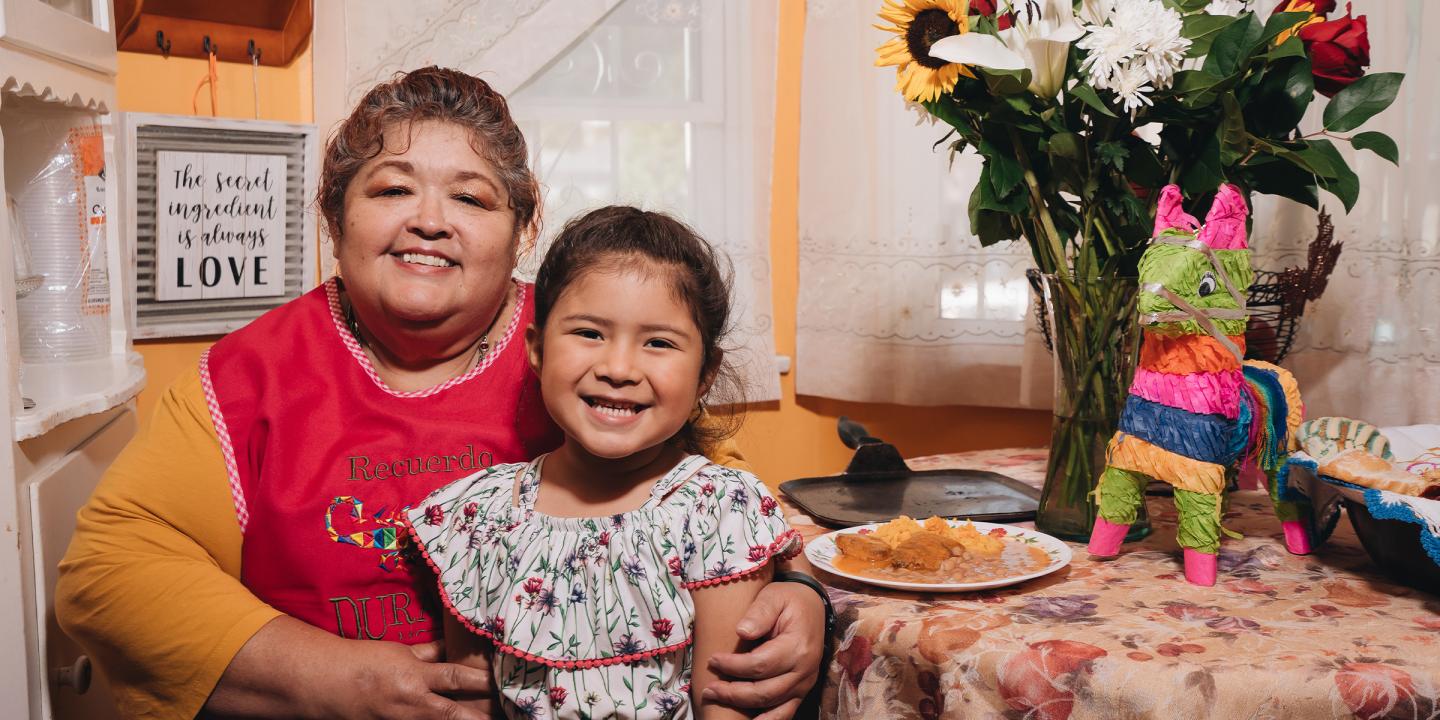 Photo of a woman and girl sitting together at a colorfully decorated table. They are both smiling at the camera. On the table are plates of food, a vase of colorful flowers, and a piñata.