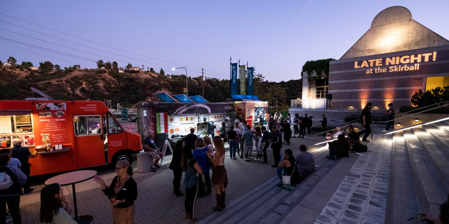 Photo of the front steps of the Skirball. People are standing around talking and ordering food from parked trucks. The words "Late Night!" are projected in lights on the side of the building.