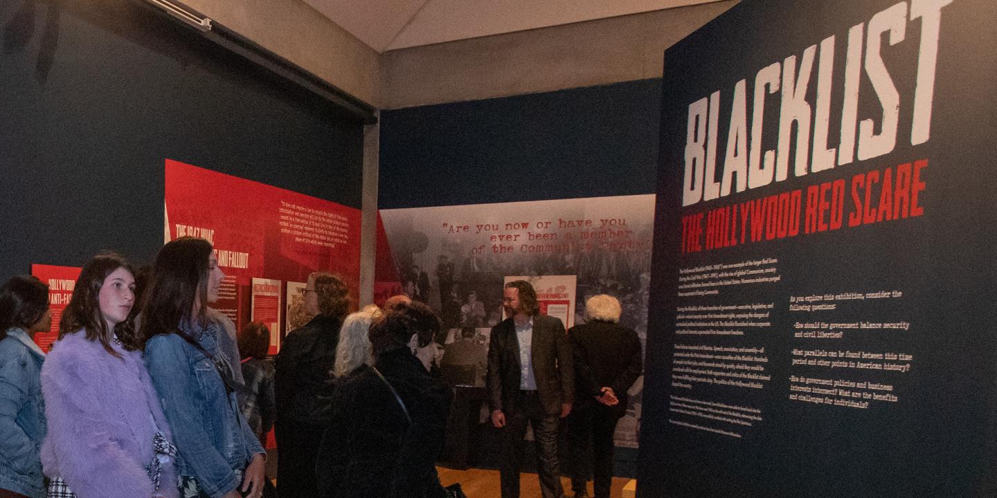Photo of a group of people entering the gallery space of Blacklist: The Hollywood Red Scare. The title can be seen on a wall to the left.