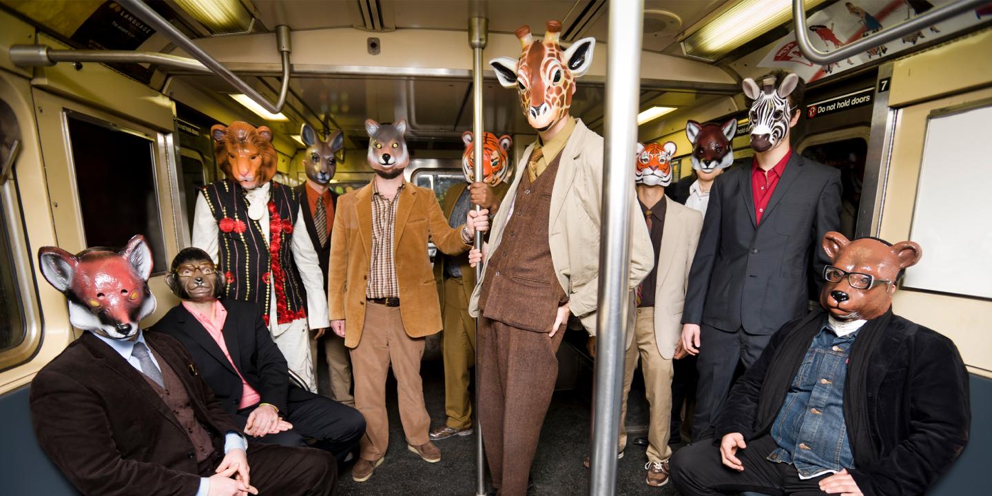 Photograph of the 11 band members sitting and standing in a subway car. They are all wearing suits and animals masks and looking at the camera.