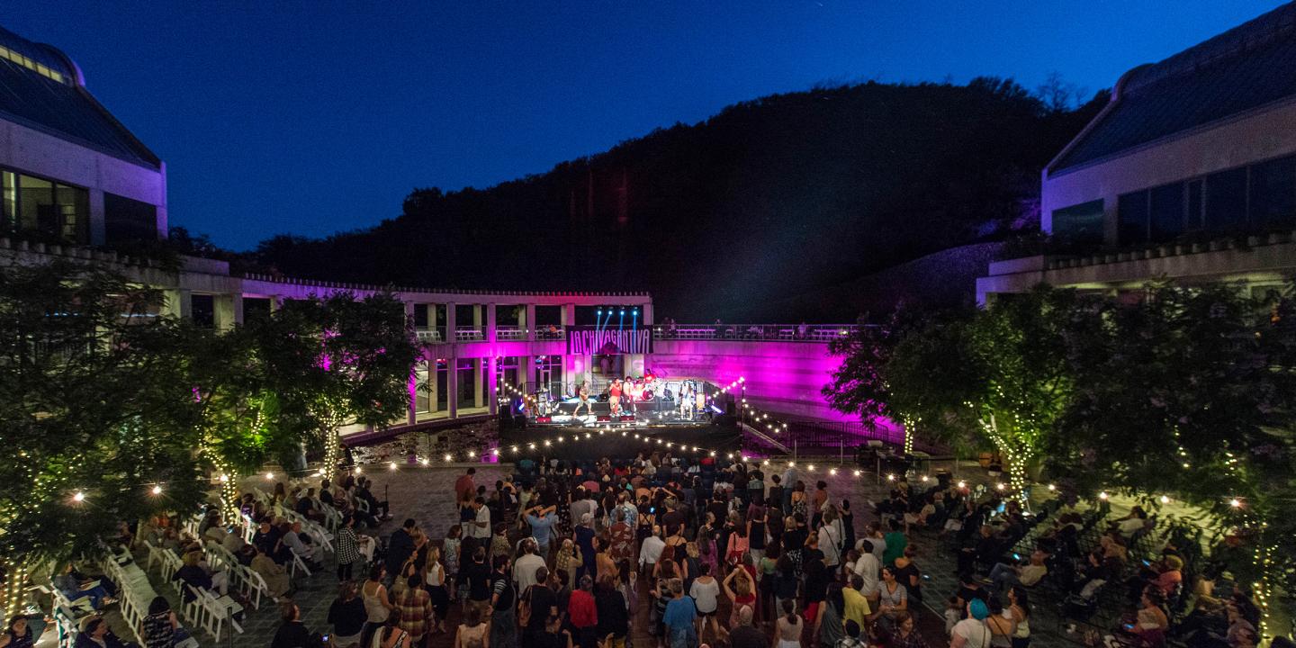 Photo of an outdoor concert at the Skirball. A large group of people are in the courtyard dancing to a band on stage. Lights are seen in the trees, strung across the courtyard, and from the stage.