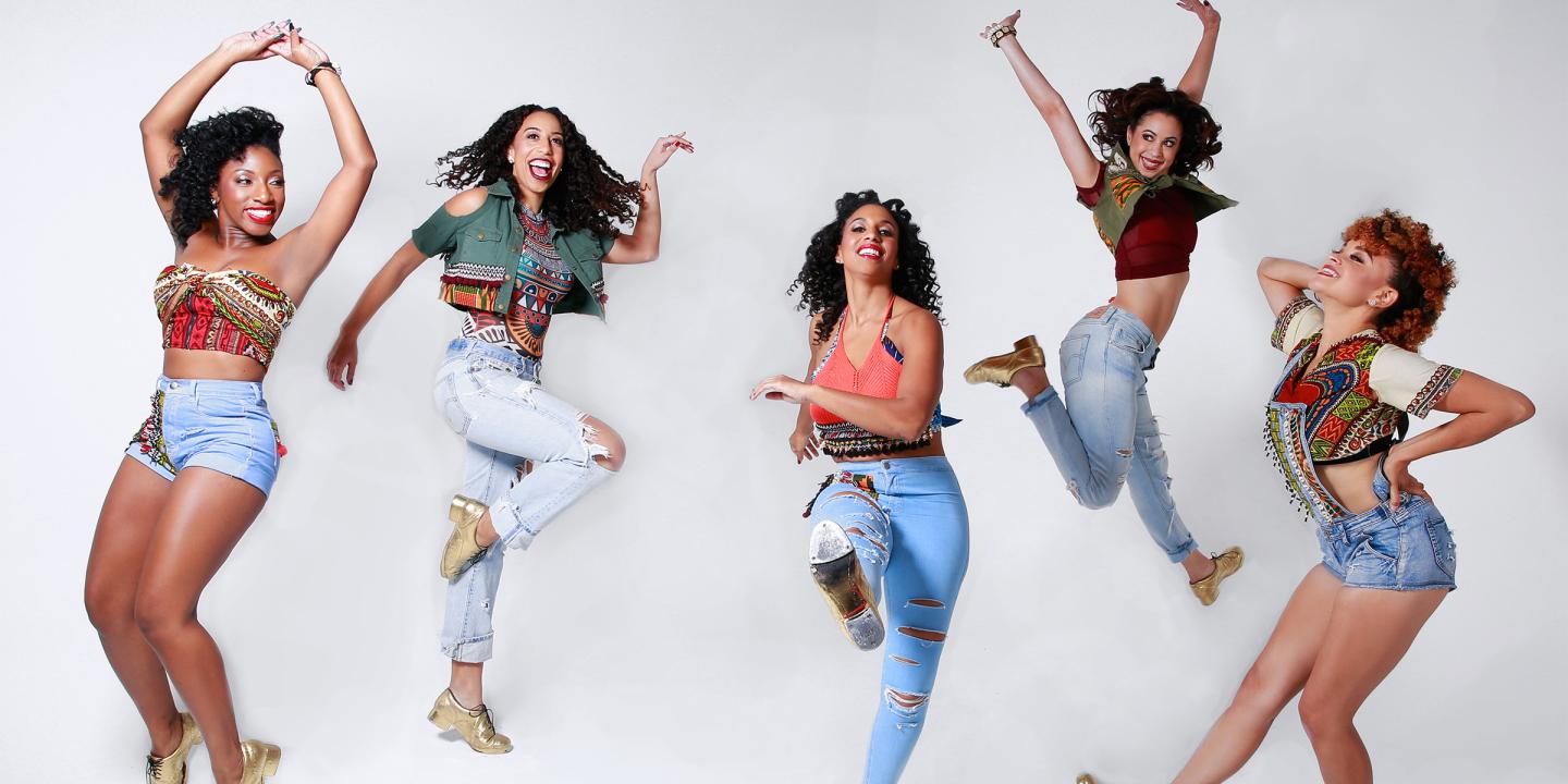 Photo of five ladies smiling and dancing in front of a white background. They are wearing colorful tops, jeans, and gold shoes.