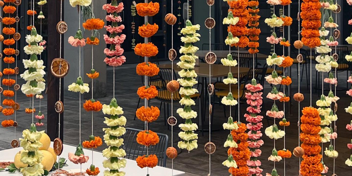 Red, yellow, and orange strands of carnations and mums hanging from a wooden structure with strings. A table and chairs with lemons on the table are in the center of the structure.