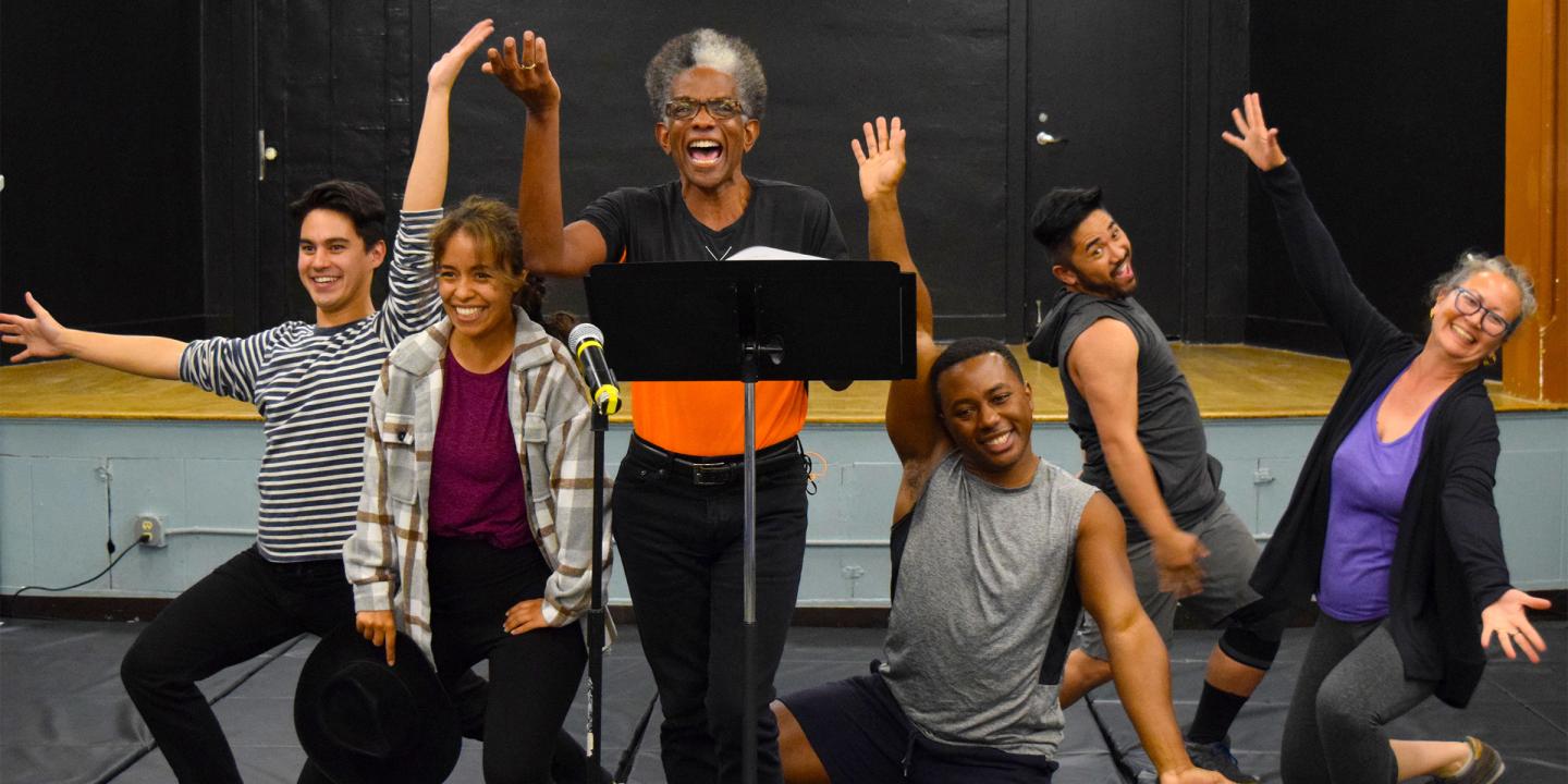 A group of six performers stand in a line posing in song facing foward.