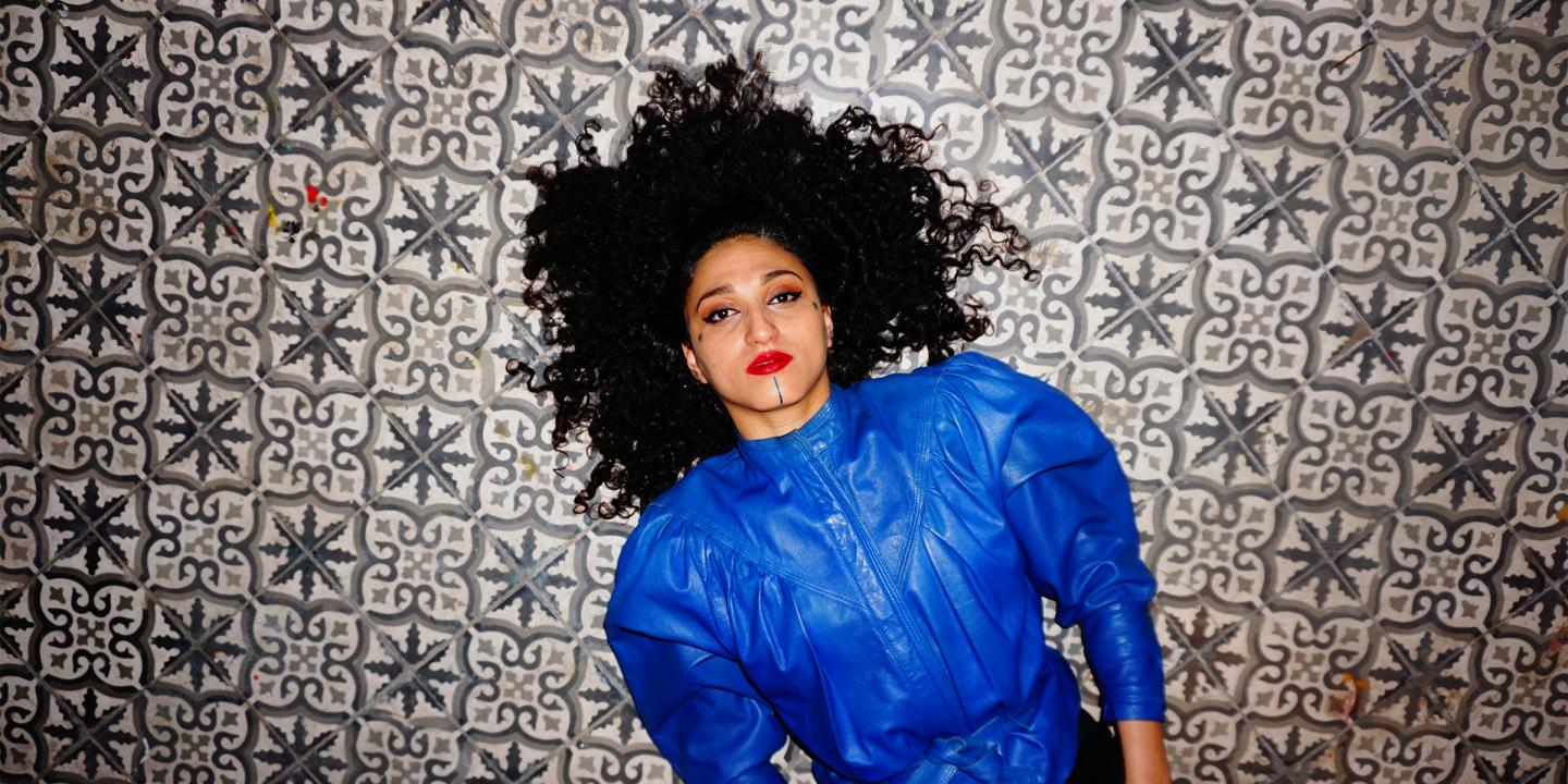 Yousra Mansour, lead singer of the band Bab L'Bluz, lies on a decorative tiled floor wearing a bright blue top. Her hair fans out around her head as she looks directly to the camera.