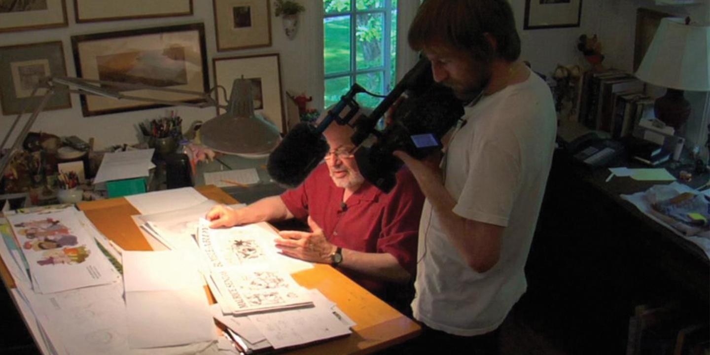 A man with a large camera stands over Maurice Sendak at his desk displaying his illustrations.