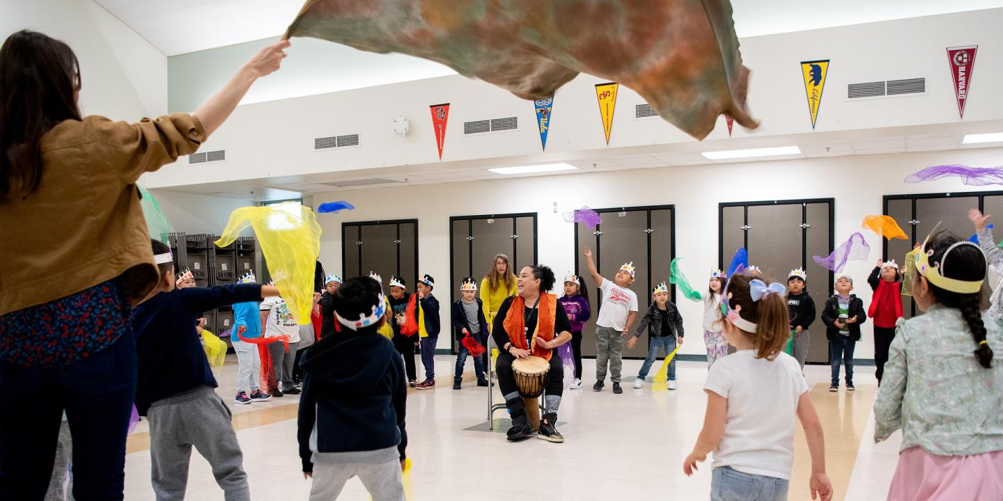 A large circle of children dancing and throwing colorful scarves while someone drums in the center.