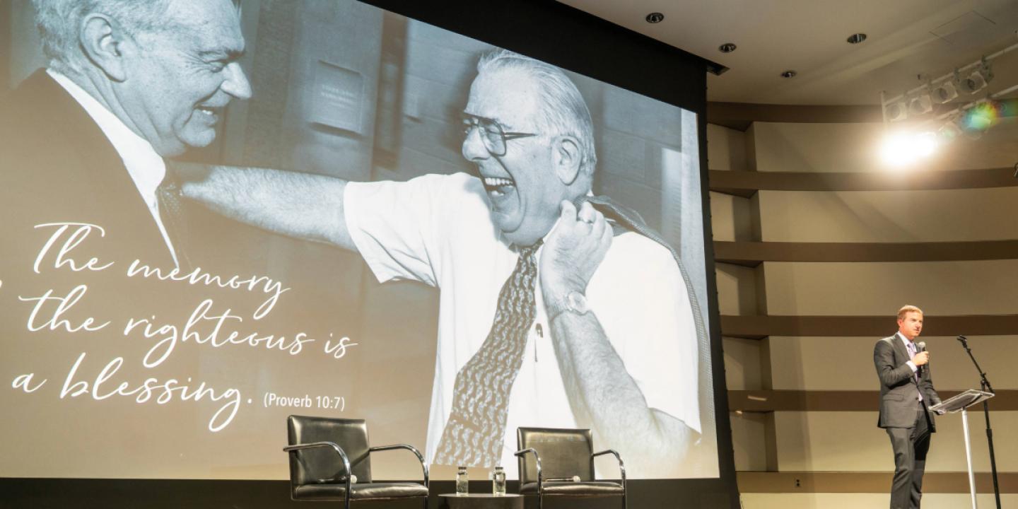 A stage is set up with two chairs on a rug with microphones for a discussion with a screen showing Uri Herscher and Howard Friedman.