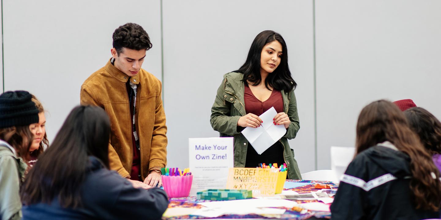 two teens standing in front of a room with other teens sitting at a table working on project.