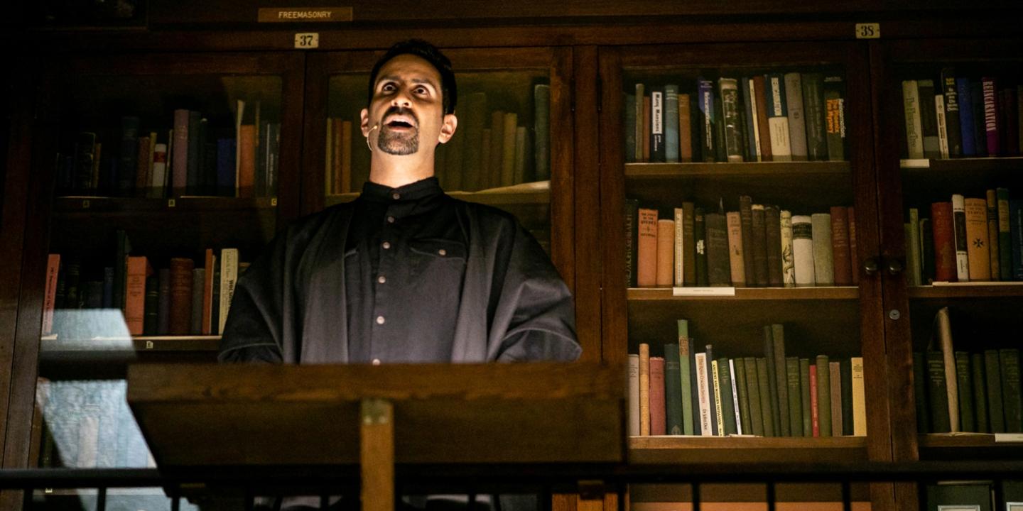 Still image of a performance featuring a man speaking in front of a tall bookcases filled with old, hardbound books.