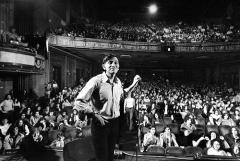 Bill Graham on stage holding a microphone stand with a crowd of people in the seats below the stage