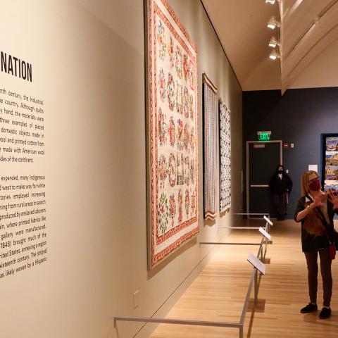 Image from inside a gallery space in Fabric of a Nation: American Quilt Stories. Large quilts are hanging on all walls and three women stand in the center of the frame discussing a quilt in front of them.