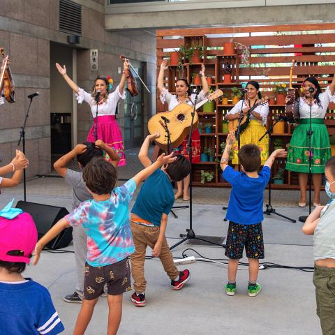 Kids dancing and watching a band of six women playing guitar and wearing brightly colored skirts