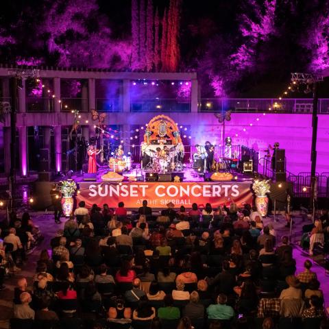 Wide view of a  nighttime concert with performers playing and a large seated audience watching. A big stage banner reads Sunset Concerts at the Skirball.