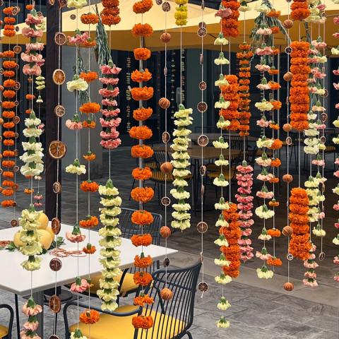 Red, yellow, and orange strands of carnations and mums hanging from a wooden structure with strings. A table and chairs with lemons on the table are in the center of the structure.