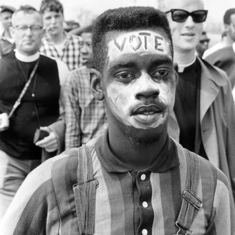Black-and-white photo of a young, African-American boy, whose face is painted white with the word "VOTE" written across his forehead
