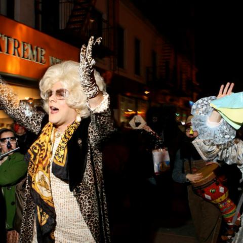 Amichai Lau-Lavie dressed as his drag character Rebbetzin Hadassah Gross dancing down a street at night with a crowd of musicians and party-goers.