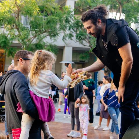 During a crowded celebration, a young child is held up by their adult to touch a tambourine in the shape of the Star of David that’s being held by a musician on a stage.