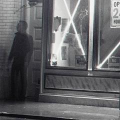 Black and white photo of shadowy man standing outside the doorway of a store at night