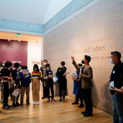 Photo of a group of students gathered around the entrance to the exhibition Visions and Values. An adult is seen gesturing to the students to orient them to the gallery.