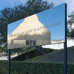 A large, mirrored sign with the words, How much of love is attention? written on it. The Skirball can be seen in the reflection.