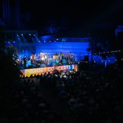 An outdoor stage in lights with performers and a crowd of people watching them